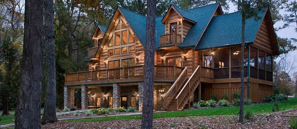 Gold tones three level log home steep gabled black roof large deck screened porch glowing windows at dusk in woods.